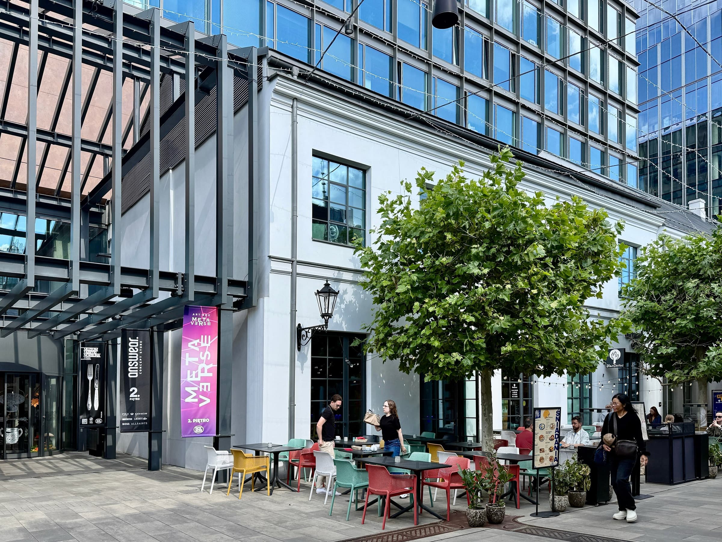 Café terrace under trees beside a restored white factory building at Fabryka Norblina